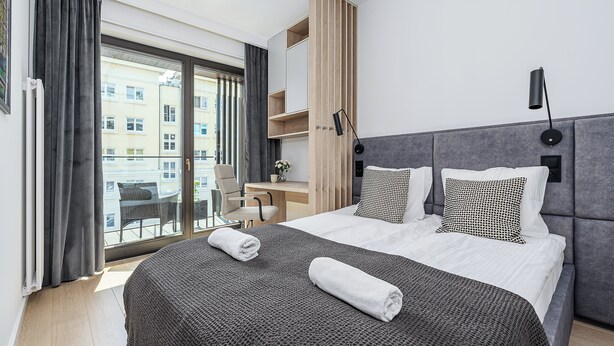 Moderni hotellihuone, jossa on suuri sänky, työpöytä ja parveke, jolta on näkymä kaupunkiin.