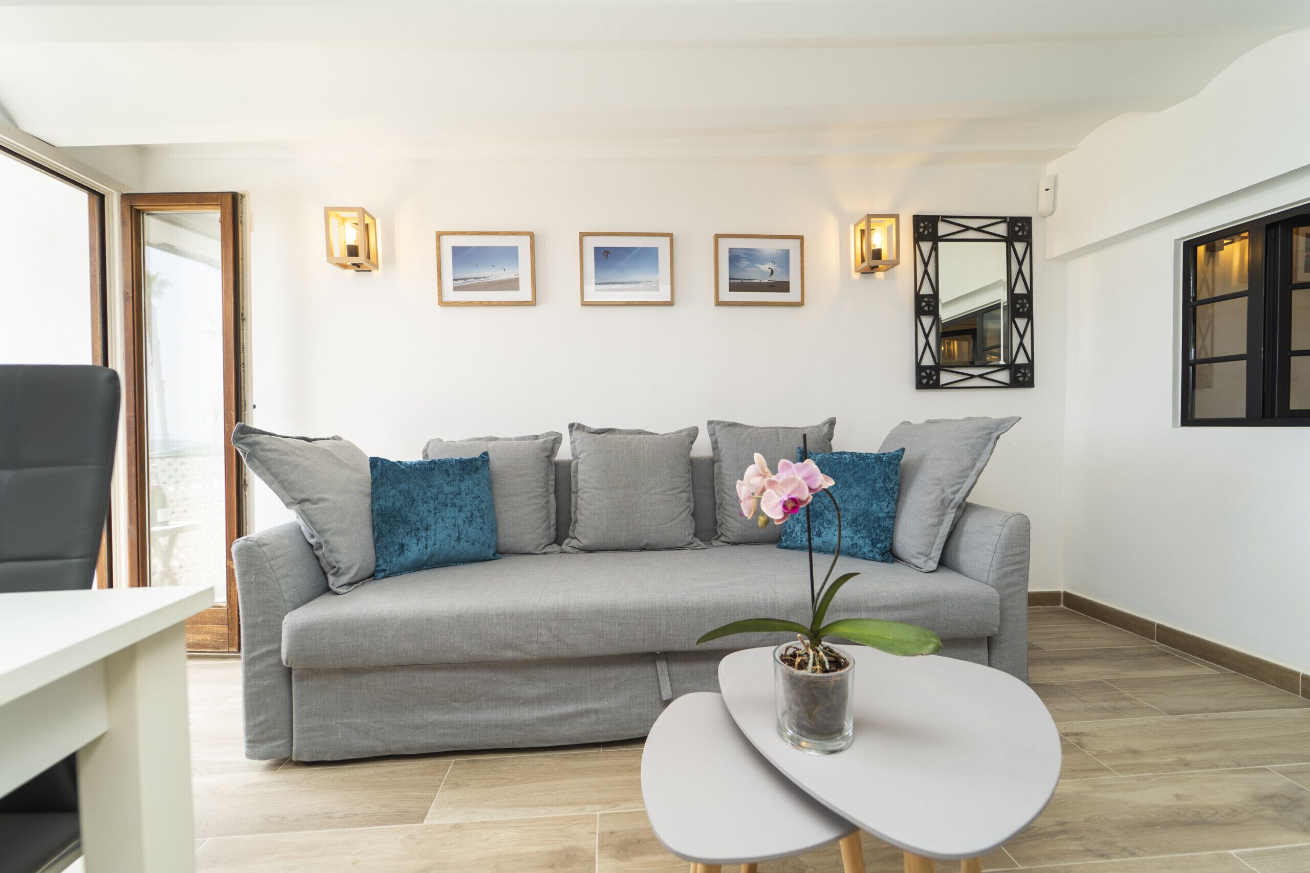 Ein modernes Wohnzimmer mit einem grauen Sofa, einem Couchtisch und einer Topf-Orchidee. An der Wand befinden sich gerahmte Bilder und ein Spiegel über dem Sofa.