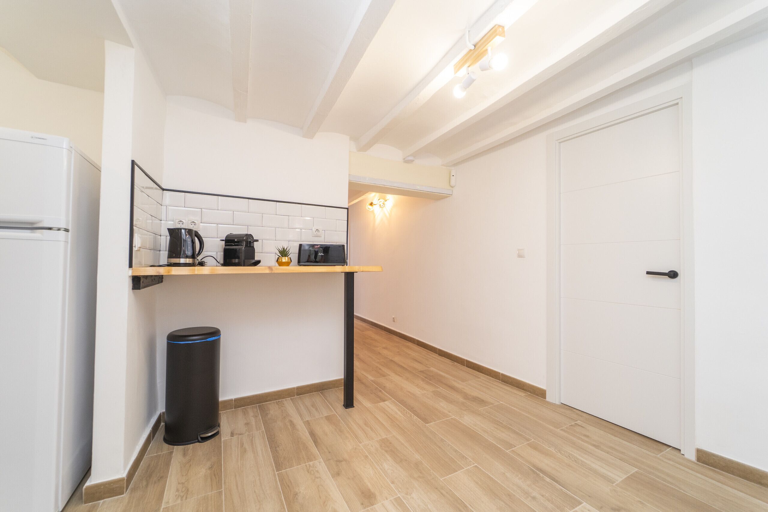 Una cocina compacta con un refrigerador blanco, una cafetera, un basurero y un estante de madera.