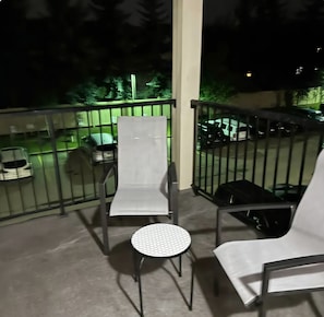 A balcony with a metal railing, a chair, a small round table, and a view of parked cars and greenery.