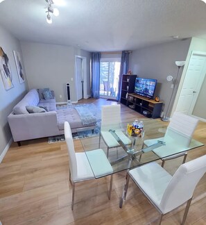 A modern living room with a glass dining table, white chairs, a sofa, and a TV.