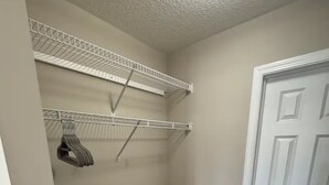 Un cuarto pequeño y cerrado con una puerta blanca, un cubo de basura gris y una estantería metálica con ropa colgada.