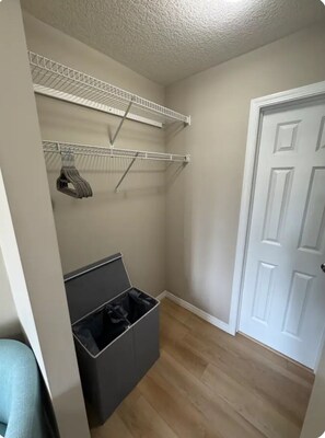 A small, enclosed room with a white door, a grey trash bin, and a metal shelving unit with clothes hanging.
