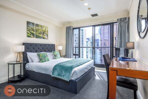 A hotel room with a large bed, a desk, and a view of the city through large windows. - Queen Street City View Studio with AC, Pool n Gym (Auckland)
