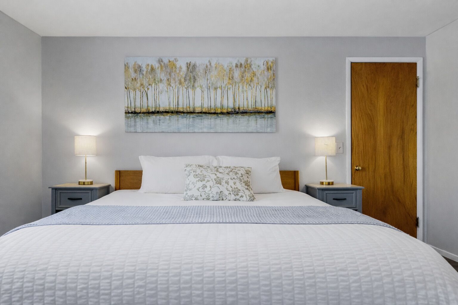 Quarto com cama grande, duas mesas de cabeceira com abajures, porta de madeira e um quadro de árvores na parede.