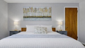 A bedroom with a large bed, two bedside tables with lamps, a wooden door, and a wall-mounted painting of trees.