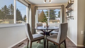 A dining area with a round table and four chairs, large windows with a view of trees and houses, and a wall-mounted shelf with decorative items.
