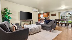 Ein modernes Wohnzimmer mit Flachbildfernseher, grauen Sofas, einem gemusterten Hocker und einem Essbereich mit Holzsesseln und einem Tisch.