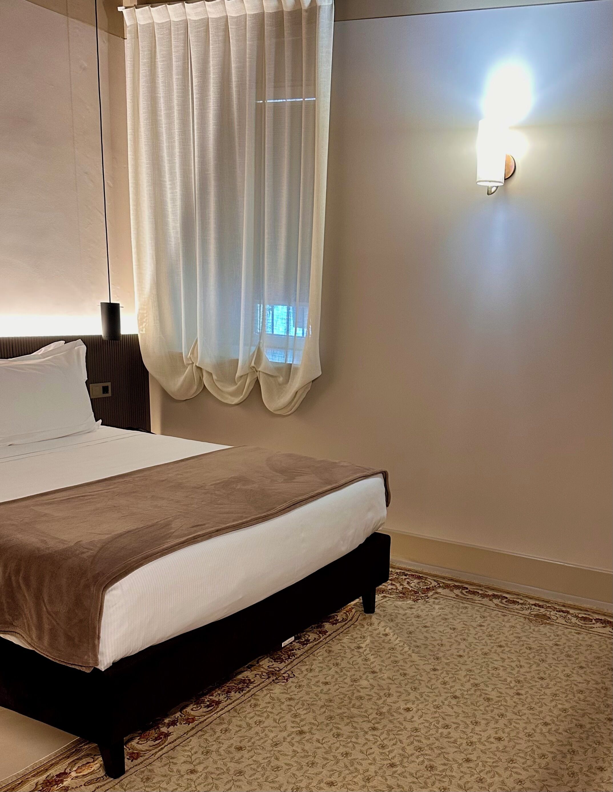 Un lit bien fait, avec une tête de lit marron, un rideau blanc et un luminaire fixé au mur.