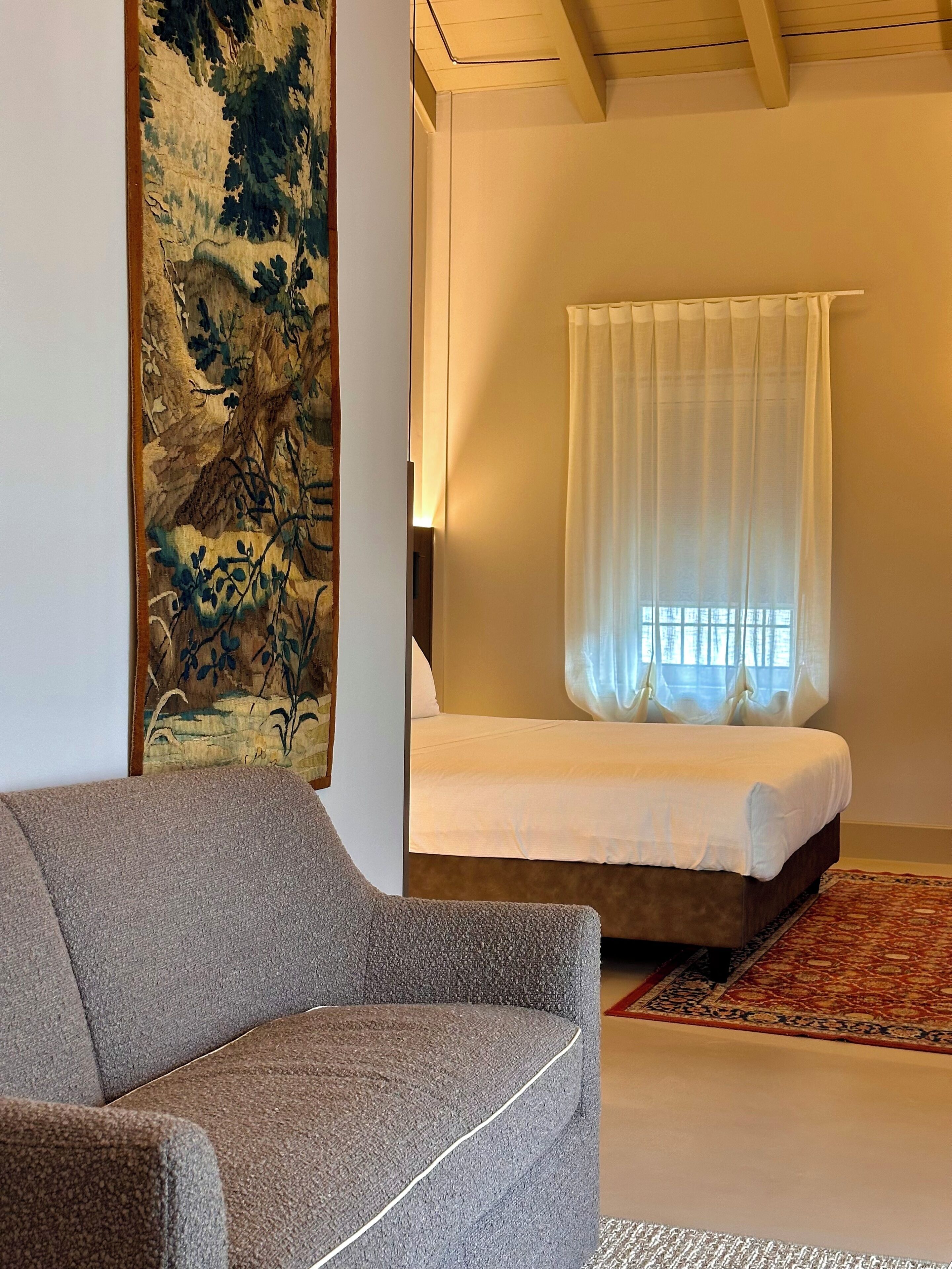 A bedroom with a bed, a large tapestry, a grey armchair, and a patterned rug.