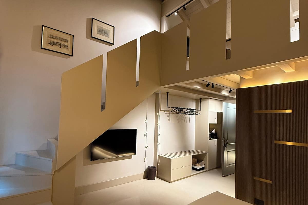A modern bedroom with a large bed, a built-in TV, a small seating area, and a staircase leading to an upper level.