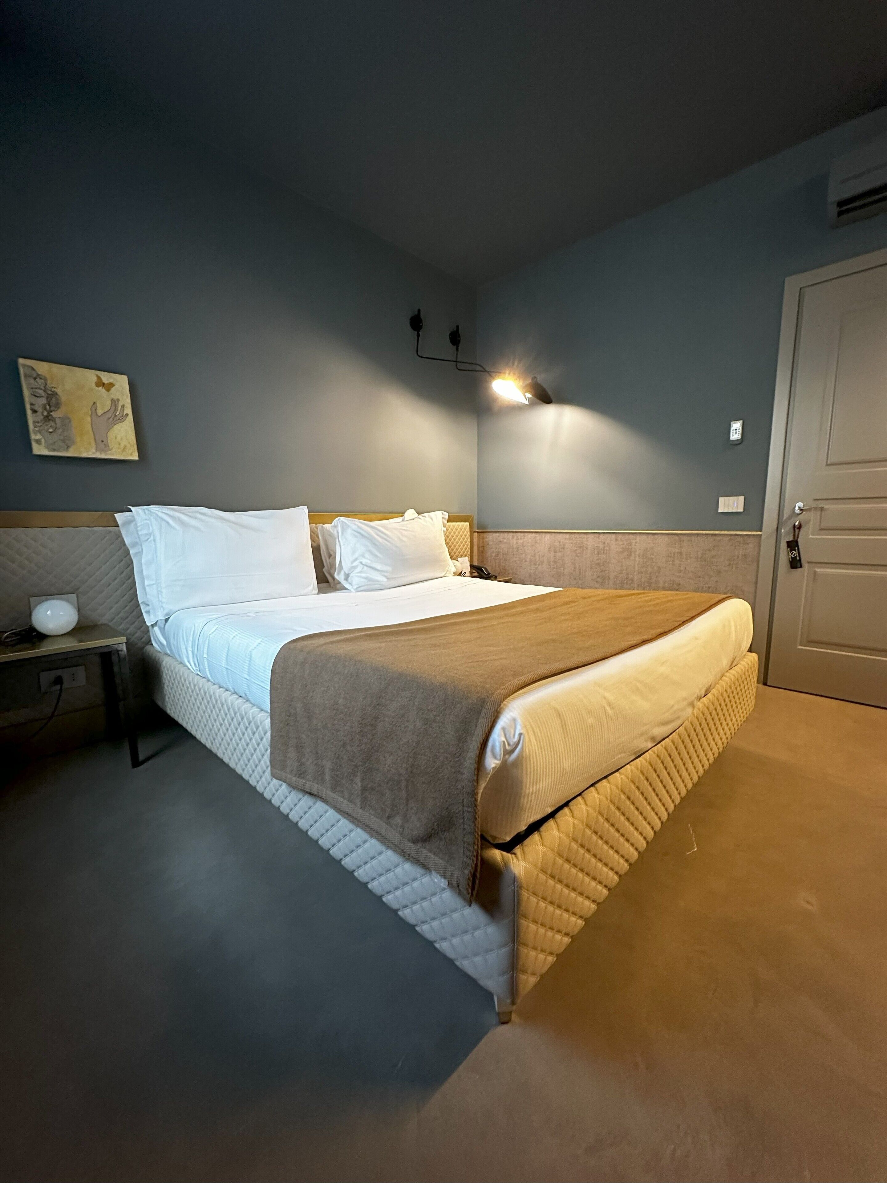 Een net opgemaakt bed met hoofdbord, een nachtkastje met een lamp en een wandlamp.