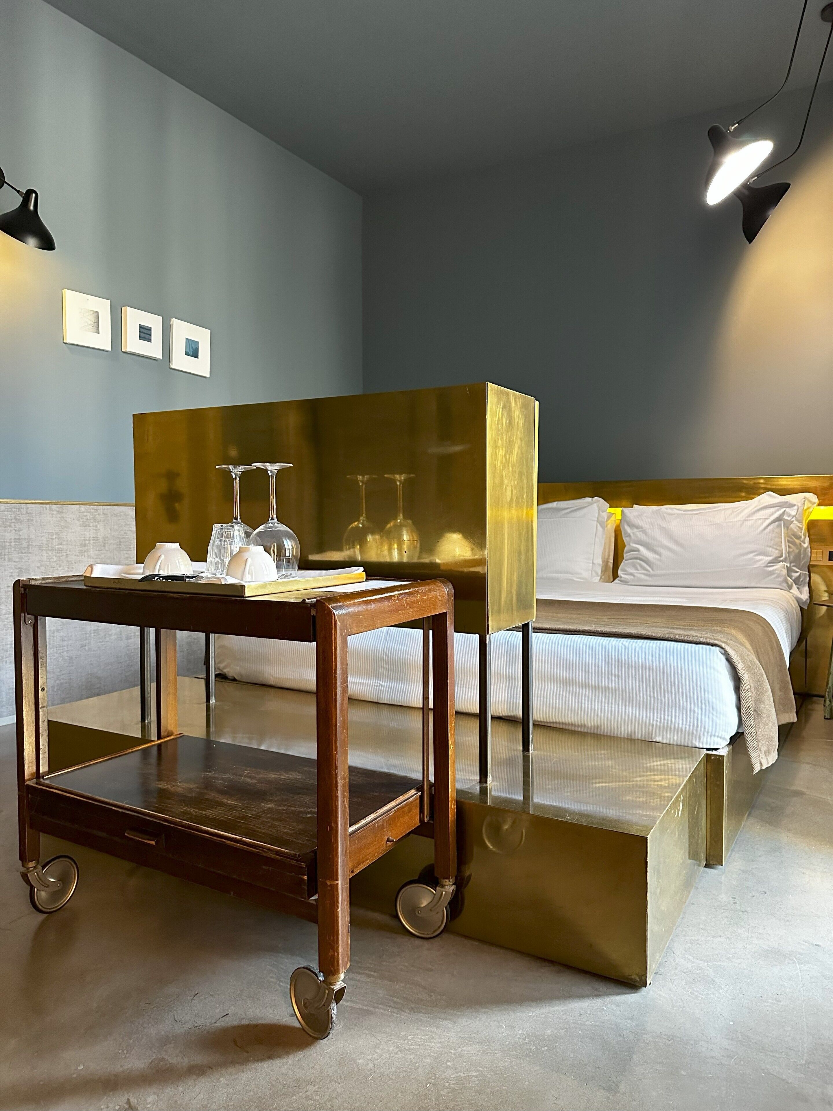 A hotel room with a bed, a wooden side table, a mirror, and a wall-mounted lamp.