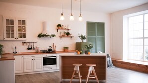 A modern kitchen with white cabinets, wooden countertops, and a central island with stools. There are hanging lights, shelves with plants, and a window with blinds.
