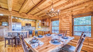 A log cabin interior with a dining table set for four, wooden cabinets, a kitchen area, and large windows. - Rustic Retreat - view-pets-games-hot tub-fire pit (Mineral Bluff)