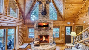 A log cabin interior with a stone fireplace, wooden walls, and a large window overlooking the outdoors. - Rustic Retreat - view-pets-games-hot tub-fire pit (Mineral Bluff)