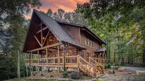 A log cabin with a porch, a staircase, and a view of the surrounding forest. - Rustic Retreat - view-pets-games-hot tub-fire pit (Mineral Bluff)