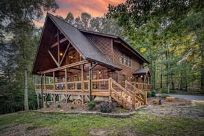 A log cabin with a porch, a staircase, and a view of the surrounding forest. - Rustic Retreat - view-pets-games-hot tub-fire pit (Mineral Bluff)