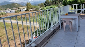 A balcony with a white railing, a table, and chairs. There is a view of a parking area and greenery in the background. - T3, in house 3 min from the beach, 5mn from the village on foot, very quiet. (Saint-Florent)