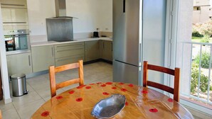 A compact kitchen with a stainless steel refrigerator, a built-in oven, and a gas stove. There is a dining table with chairs, and a trash bin is visible. - T3, in house 3 min from the beach, 5mn from the village on foot, very quiet. (Saint-Florent)