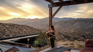 A person sitting on a wooden deck with a view of mountains, a dog nearby, and outdoor furniture. - Hawkeye Dome • 112 Acres, Pool & Spa, Epic Views (Yucca Valley)