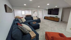 Ein Wohnzimmer mit einer Eckcouch, einem Hocker und einem Fernseher auf einem Standfuß.