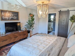 Ein Schlafzimmer mit einem großen Bett, einem Fernseher auf einer Holzkommode, einer Kristallleuchtre und einer Schiebetür zu einem Badezimmer.