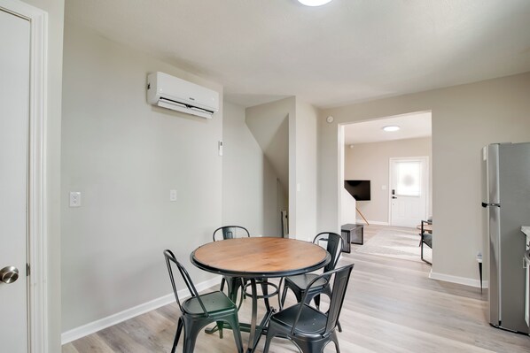 Un comedor con una mesa redonda y sillas, un equipo de aire acondicionado en la pared, y una zona de cocina con un refrigerador.
