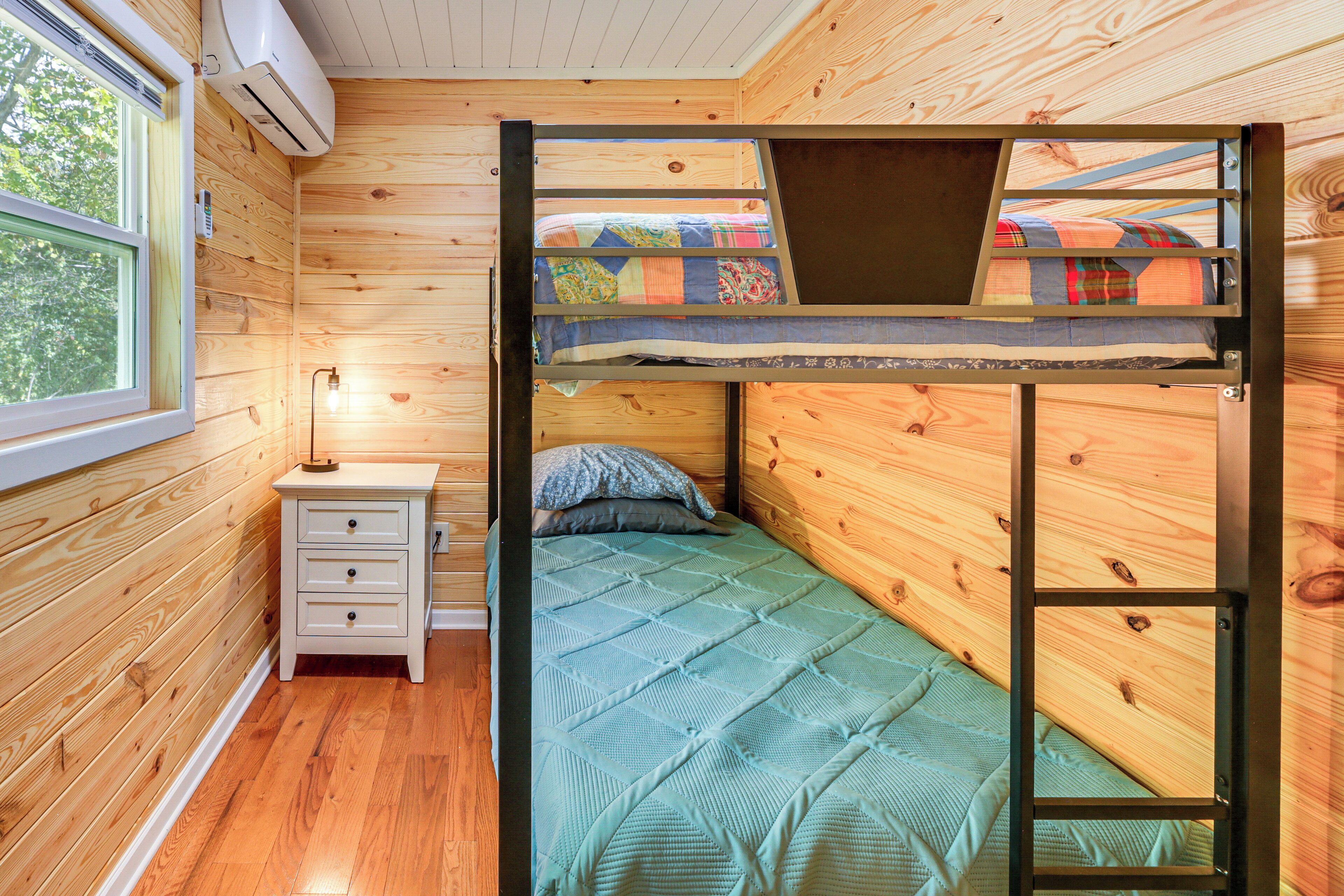 Una cama litera con colcha verde acolchada, una mesita de noche de madera con cajones y una ventana con vistas a árboles.