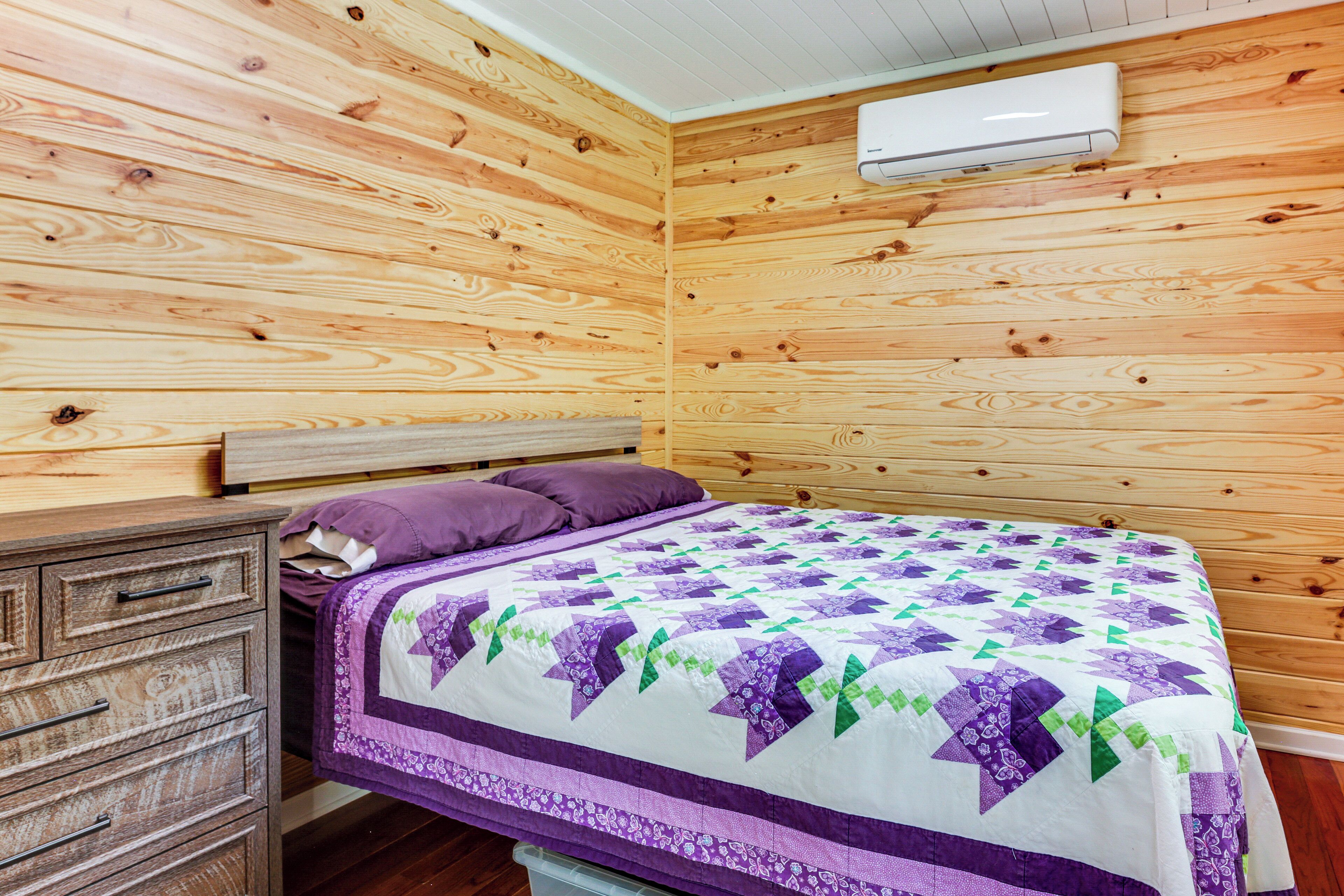A bedroom with wooden walls, a bed with a purple and white quilt, a wooden nightstand, and a white air conditioning unit.