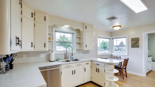 Una cocina moderna con armarios blancos, lavavajillas, un lavamanos y una zona de comedor con mesa y sillas.