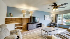 Una sala de estar moderna con chimenea de ladrillo, televisor de pantalla plana, ventilador de techo y un amplio ventanal con vistas al exterior.