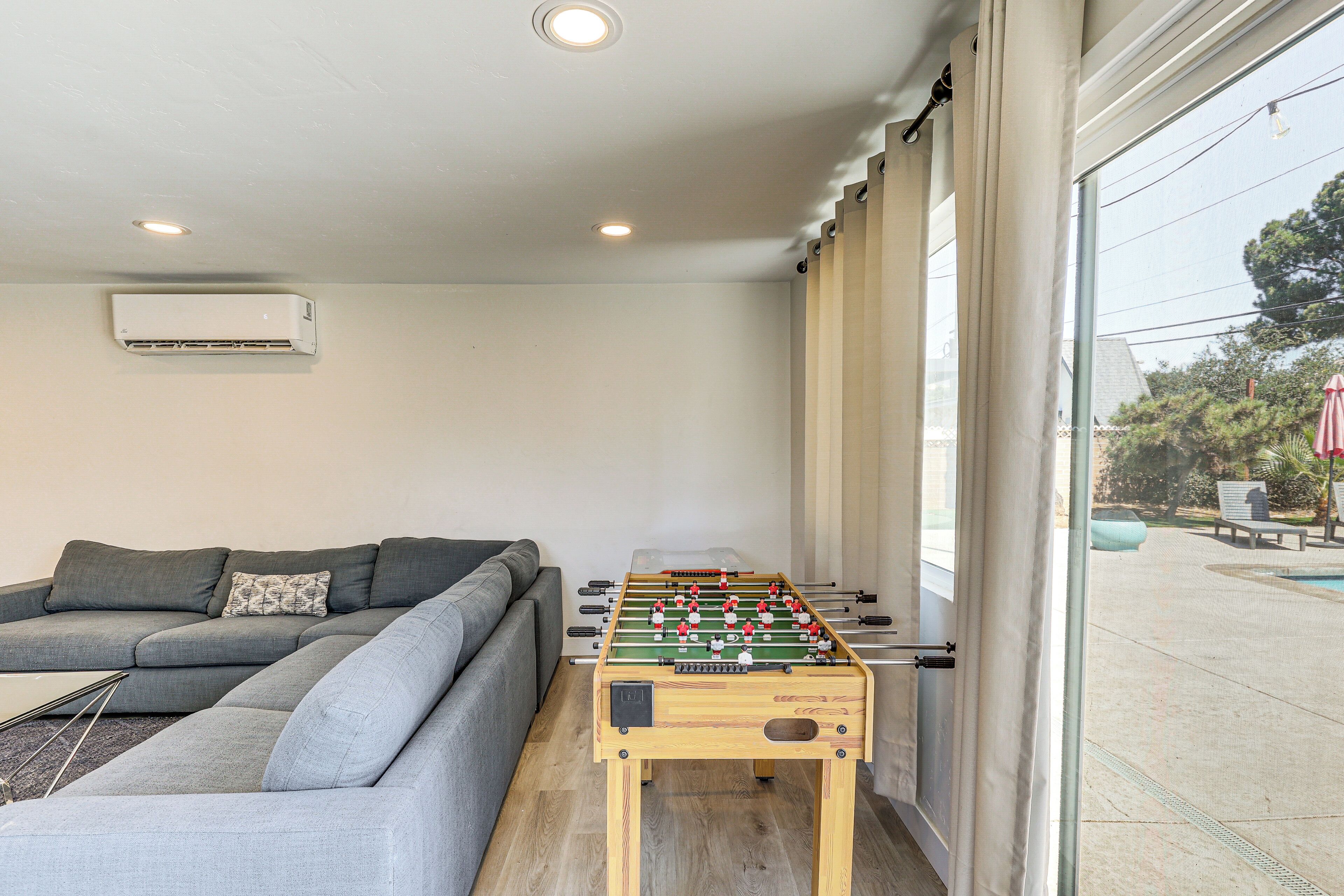 A modern living room with a foosball table, grey sofa, and a large window with curtains.