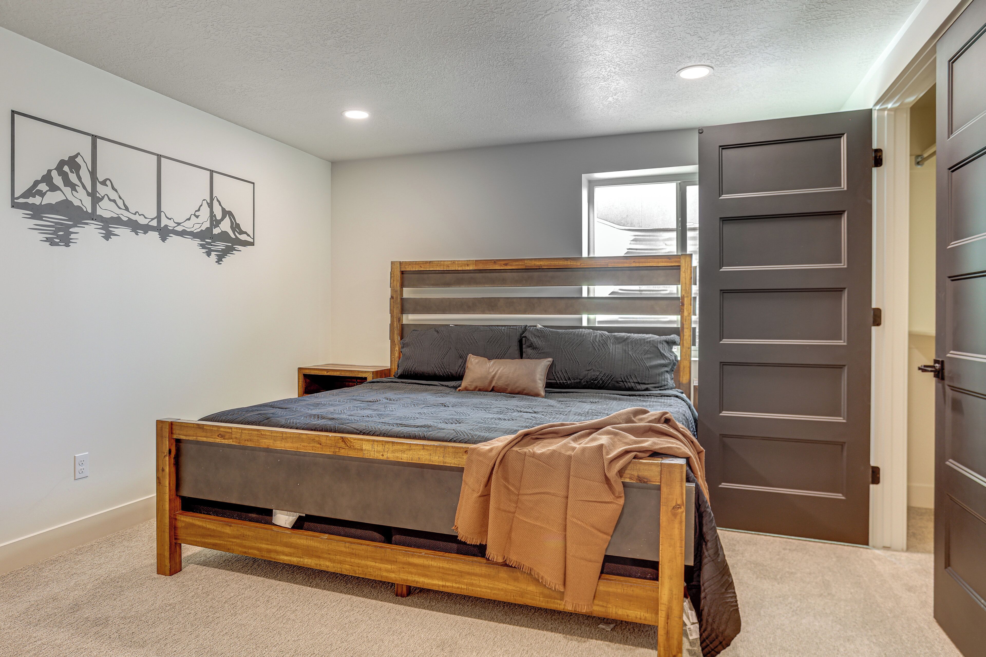 A bedroom with a wooden bed, grey bedding, a mountain artwork on the wall, and a door to the right.