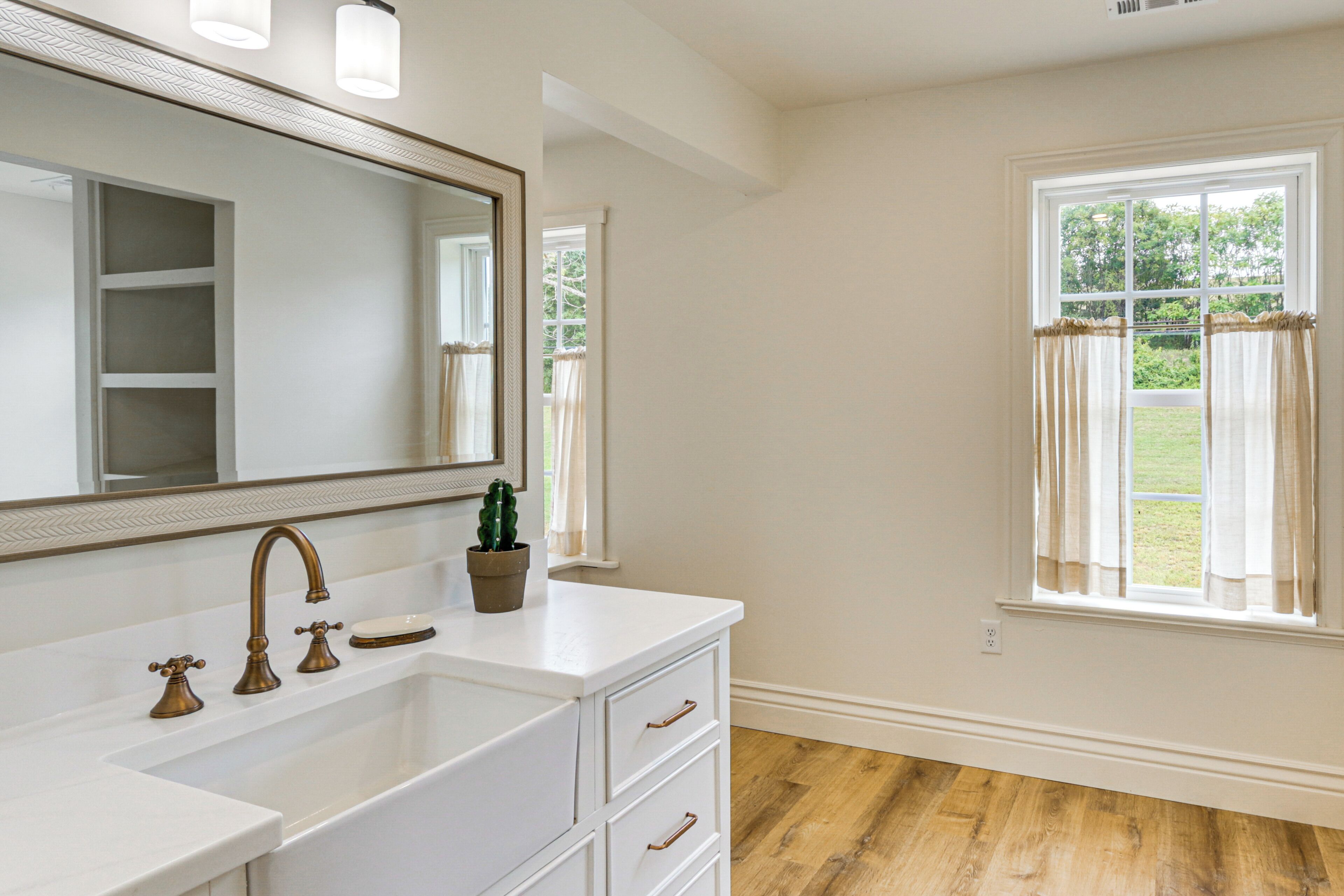 A bathroom with a white vanity, a large mirror, a sink with a single faucet, and a window with curtains.