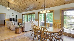 Un vaste salon doté d’un plafond en bois, un coin repas avec des chaises et une table en bois, et de grandes fenêtres offrant une vue sur l’extérieur.