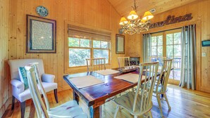 A wooden interior with a dining table, chairs, a chandelier, and large windows.