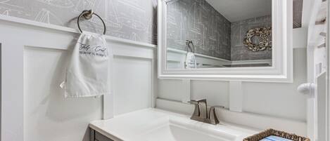 Een badkamer met een witte wastafel, een spiegel en een handdoek aan een ring. Op de muur hangt een decoratieve krans en op het aanrecht staat een klein rieten mandje.