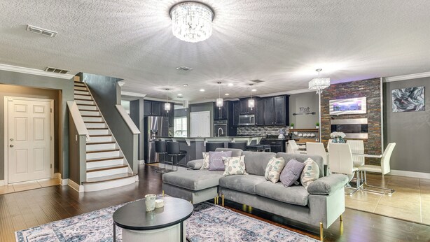 A spacious living area with a gray sectional sofa, a round coffee table, and a patterned rug. A staircase leads to another level, and a kitchen with dark cabinets and a stone accent wall is visible in the background.