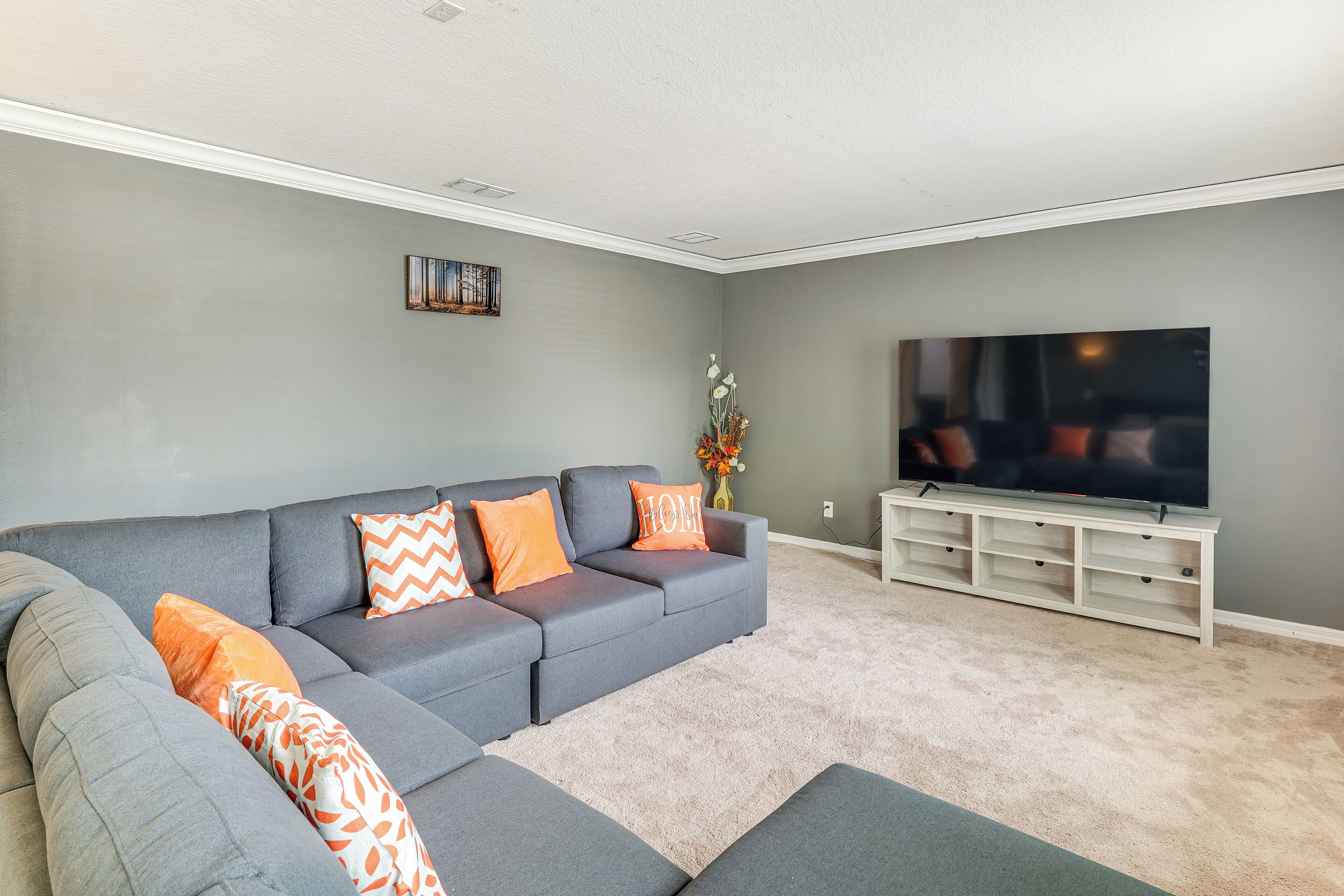 A living room with a grey sectional sofa, orange patterned pillows, a flat-screen TV on a white stand, and a beige carpet.