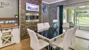 A modern dining area with a glass table and white chairs, a brick accent wall, a mounted TV, and a view of the outdoors.