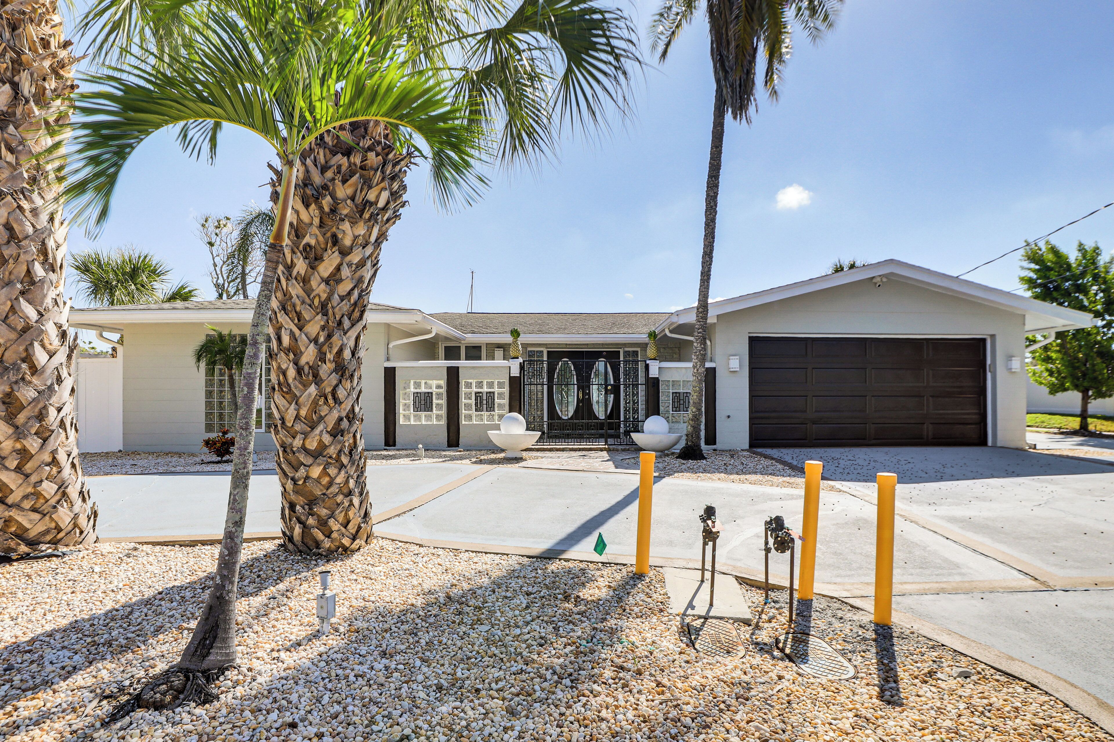 A two-story house with a garage, surrounded by palm trees and a gravel driveway.