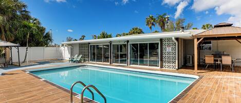 A swimming pool with a wooden deck, glass enclosure, and palm trees in the background.