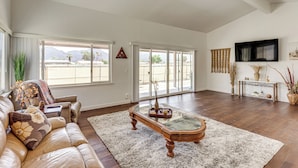 A spacious living room with a large window, a sofa, a coffee table, and a television mounted on the wall. - Mtn-view Getaway w/ Pool: 4 Mi to Dtwn Palm Desert (Palm Desert)