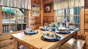 Ein Holzblockhaus-Innenraum mit einem für vier Personen gedeckten Esstisch, zwei mit Vorhängen versehenen Fenstern, einem gerahmten Bild und Blick auf ein grünes Gebäude im Freien.
