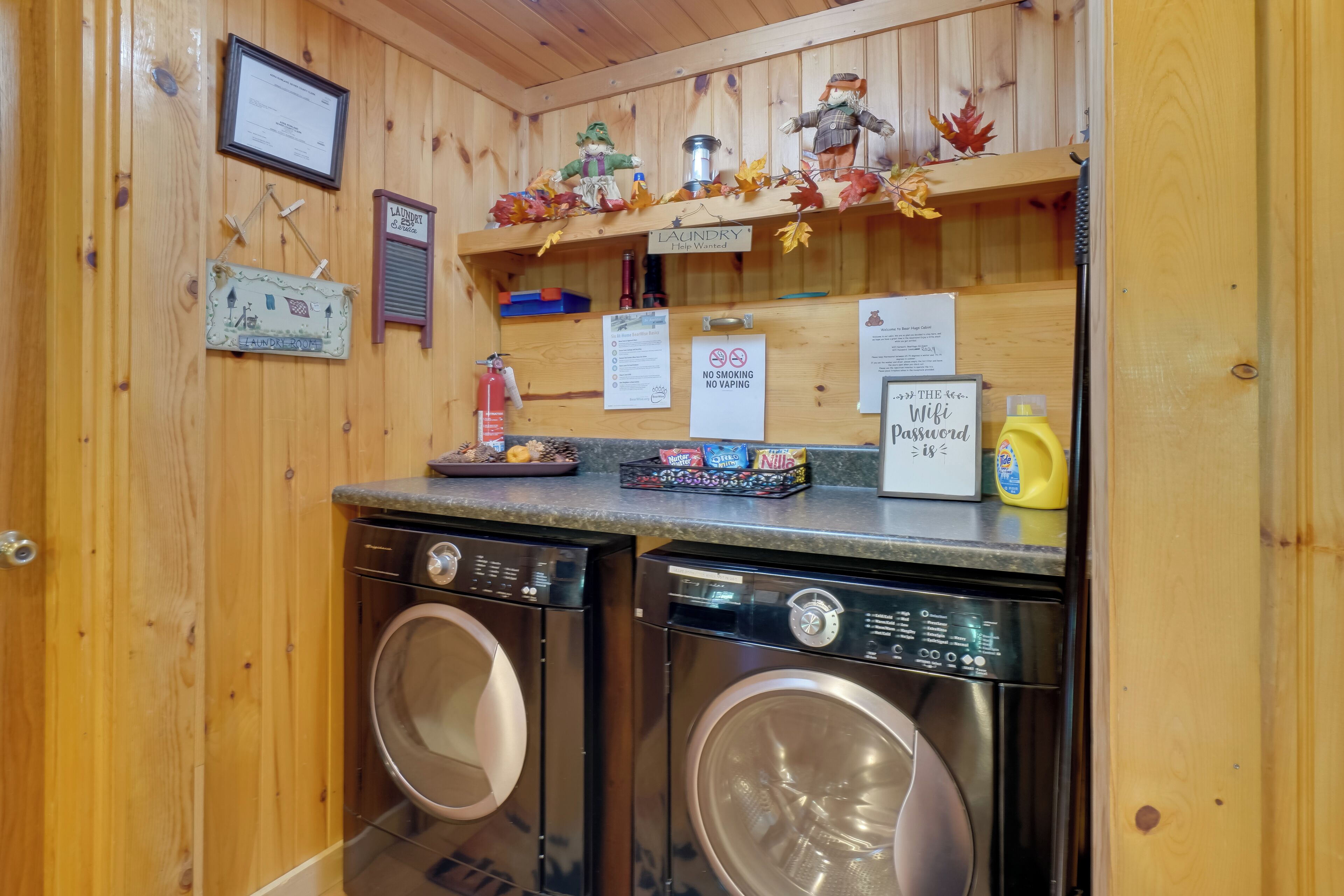 A laundry area with two front-loading washing machines, a shelf with decorative items, and signs indicating no smoking and no vaping.