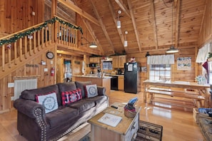 A cozy cabin interior with a wooden staircase, a living room with a leather couch, a kitchen area, and a dining space.
