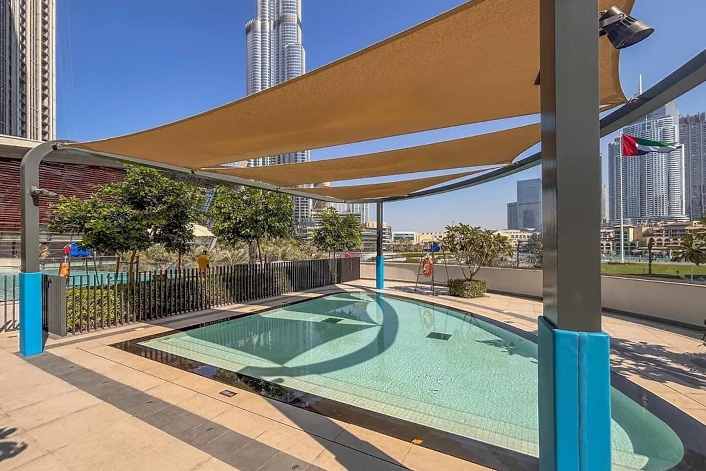 A rooftop pool with a view of tall buildings, surrounded by a fence and shaded by a canopy.