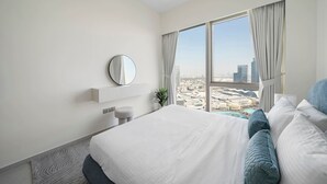 A modern hotel room with a large bed, a round mirror, a small stool, and a view of a cityscape through large windows. - Iconic Burj Khalifa & Fountain Views, 2BR Downtown (Dubai)