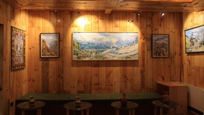 A wooden-paneled room with a large painting of a mountainous landscape, a green bench, and small round tables with candles.